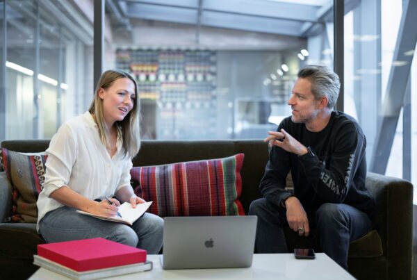 A woman and a man are sitting on a sofa in an office. They are talking and the woman is writing in a notebook. There is a laptop open on the table in front of them.